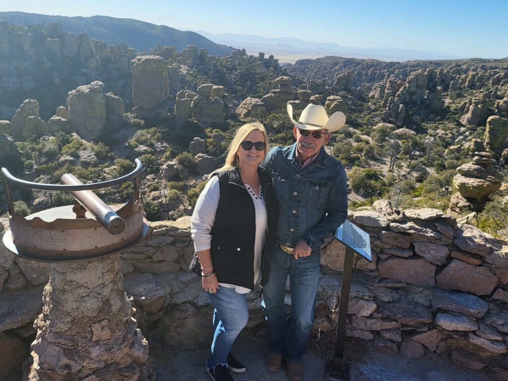 picture of a woman Lesley Mouton and a man Vincent Sanchez standing at a lookout point at the Chiricahua Monument. the woman is wearing blue jeans, white shirt and black puffer vest. The man is wearing blue jeans, a white shirt and blue jean jacket with a handmade cowboy hat