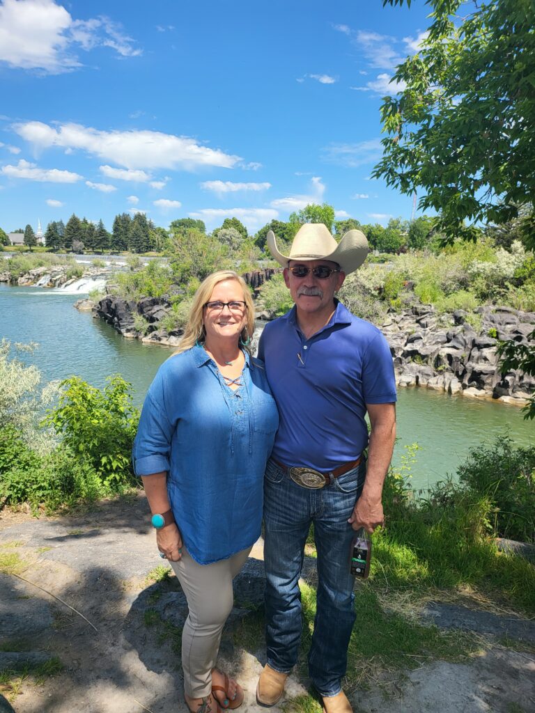 standing in front of the falls in Idaho falls, Idaho, there is a blonde woman wearing a blue jearn shirt and khaki pants with brown sandlas. The man is wearing a light blue polo styled shirt, blue jearns, cowboy boots and hat