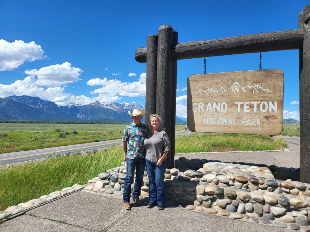 Vincent and Lesley standing under the sign to the Grand Teton National Park. Vincent is wearing a Hawaiian style button down shirt, Levi blue jeans, Paul Bonds custom boots and a belt buckle with a handmade leather tooled belt. He has on cowboy style hat. Lesley is a blonde woman wearing blue jeans, an army green shirt with black eel skin boots.