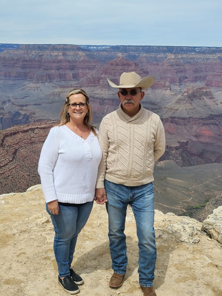 A woman and a man standing next to each other on the edge of a cliff, holding hands with their back to the Grand Canyon. The blonde woman is wearing black glasses, silver jewelry with one ring made of turquoise. She has one a white sweater with blue jeans and black shoes. The man is wearing an off white sweater with Levi blue jeans, a custom hat made by Jim Spradly, Serengeti sunglasses and Paul Bond custom boots.