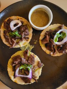 Mexico City style tacos. Small flat corn tortillas with carne asada, red onion and cilantro with a white bowl with spicy jalapeño dipping sauce.