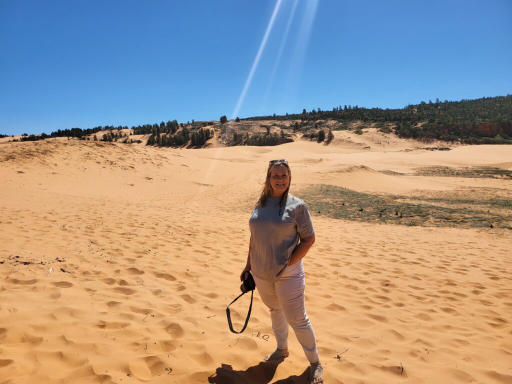 The picture is of Lesley Mouton. She is in Coral Pink Sand dunes in Kanab, UT. She's standing with her left hand in the front pocket of her white jeans and holding a Cannon EOS RP camera in her hand. She's wearing sunglasses on her head and white sandals with a River Bend Ranch tshirt on.