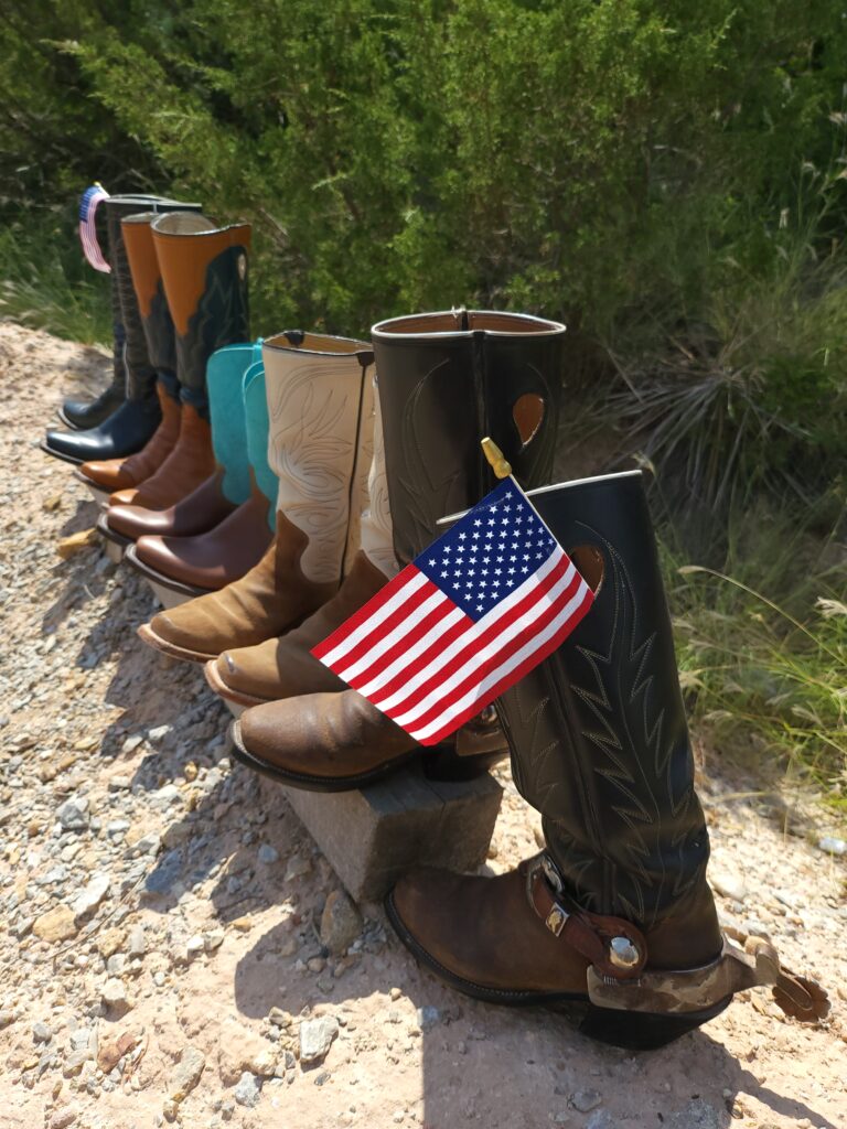a row of custom-made leather boots with American flags on either end. they are on a wooden timber on the ground in Santa Anna Texas